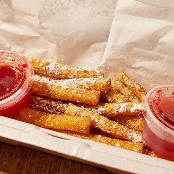 Golden churro fries dusted with powdered sugar and two cups of red dipping sauce in a takeout container.