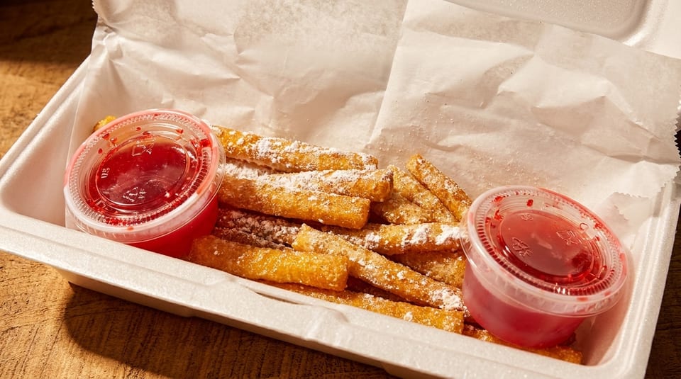 Golden churro fries dusted with powdered sugar and two cups of red dipping sauce in a takeout container.