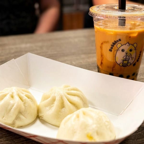 Three steamed buns in a paper tray next to a bubble tea drink on a wooden table.