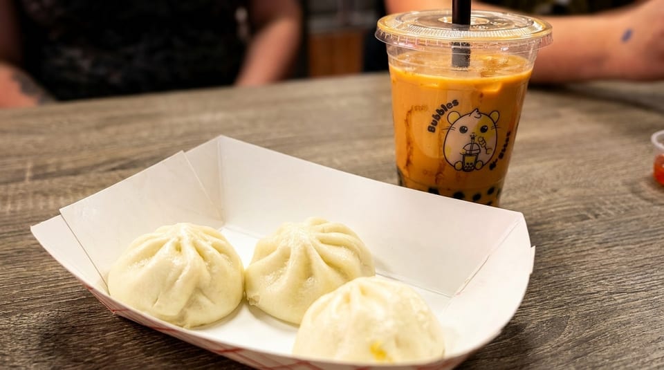 Three steamed buns in a paper tray next to a bubble tea drink on a wooden table.