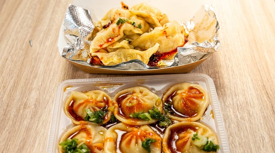 Two containers of dumplings, one with chili oil and the other with sauce and green onions.